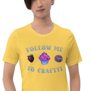 Może przedstawiać: Musztardowa koszulka z napisem "FOLLOW ME TO CRAFTY!" oraz ilustracjami truskawki, babeczki i kanapki. Koszulka wykonana jest z miękkiego materiału i nadaje się do noszenia na co dzień.
