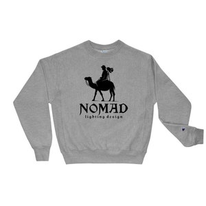 May include: A heather gray sweatshirt with a black graphic of a person riding a camel, carrying equipment. The word "NOMAD" is above the text "lighting design" in a stylized font. The Champion logo is visible on the left sleeve.