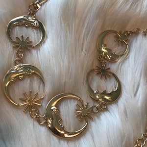 May include: Gold chain necklace with crescent moon and star charms. The chain is made of linked circles and the charms are detailed with a face on the moon.