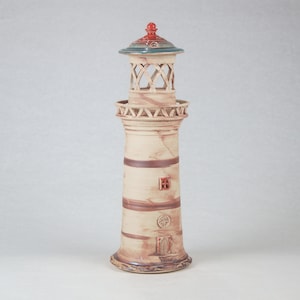 以下が含まれることがあります： 赤い屋根、緑色の頂上、茶色と白のストライプが施されたセラミック製の灯台の形をした置物。灯台には小さな窓とドアがあります。