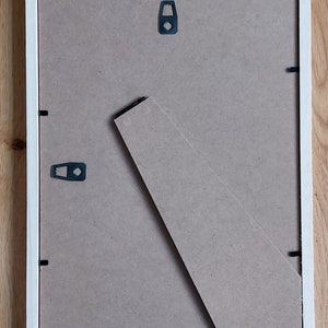 May include: The back of a white picture frame with a brown cardboard backing. The frame has a built-in easel stand and two metal hanging hooks.