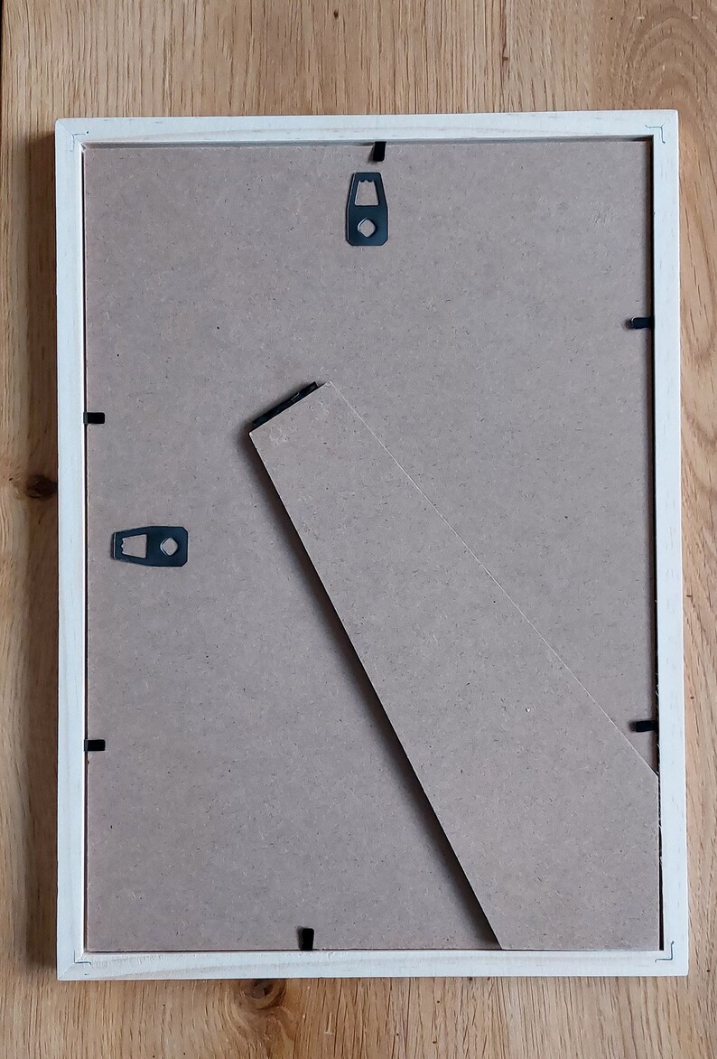 May include: The back of a white picture frame with a brown cardboard backing. The frame has a built-in easel stand and two metal hanging hooks.