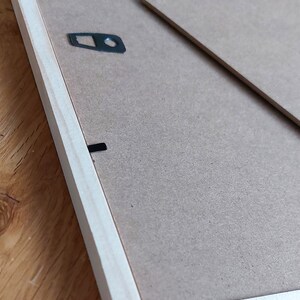 May include: The back of a light wood picture frame with a brown cardboard backing. The frame has two black metal hanging hooks and a black metal D-ring.