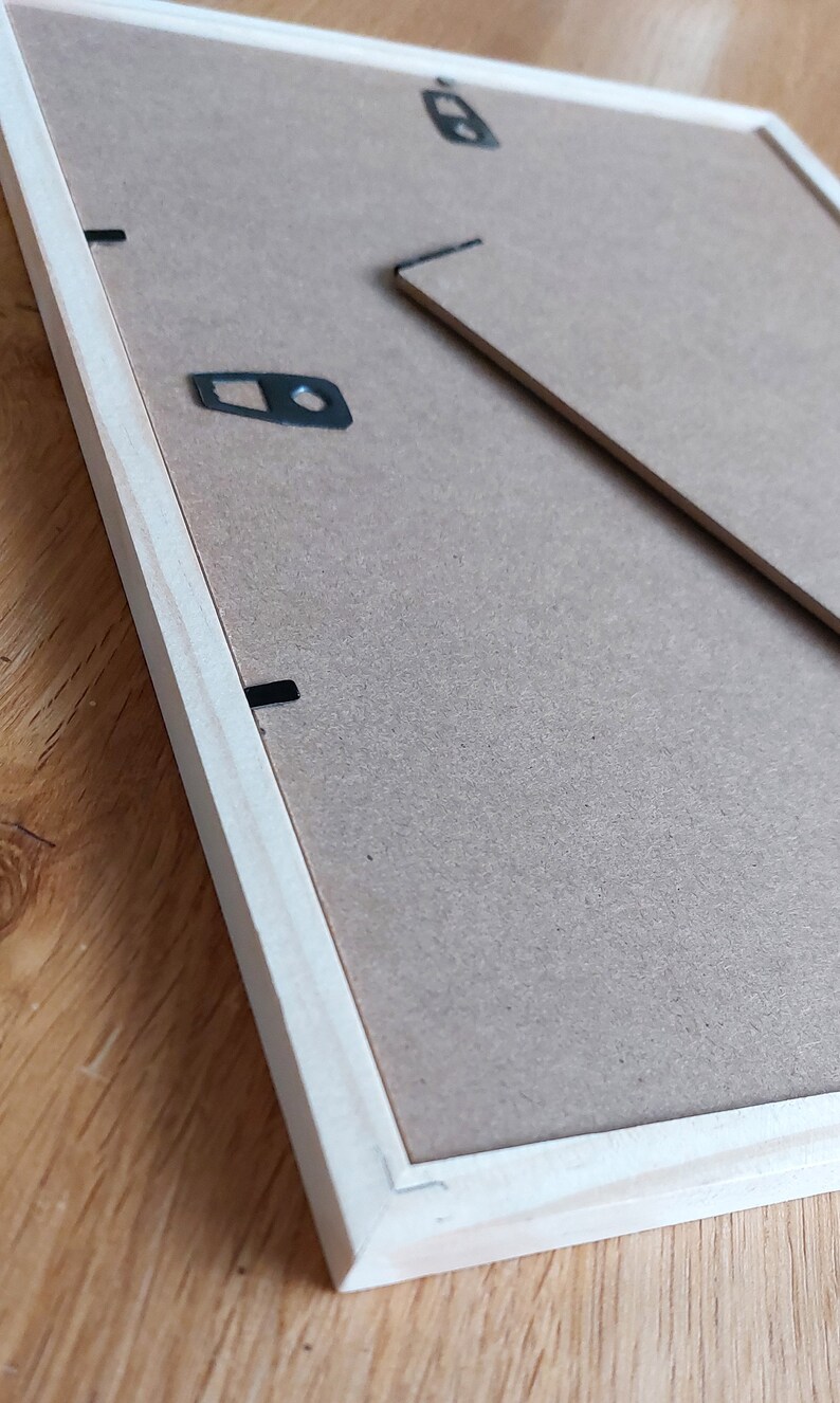May include: The back of a light wood picture frame with a brown cardboard backing. The frame has two black metal hanging hooks and a black metal D-ring.