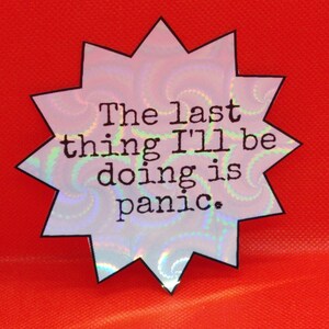 May include: A star-shaped sticker with a holographic, iridescent surface and a black outline. The text "The last thing I'll be doing is panic" is printed in black, centered on the sticker, against a bright red background.