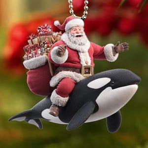Puede incluir: Un adorno navideño que representa a Papá Noel montado en una orca negra y blanca. Papá Noel viste un traje rojo y lleva un saco de regalos. El adorno cuelga de una cadena plateada sobre un fondo borroso de vegetación y bayas rojas.