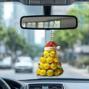 May include: A yellow pickleball Christmas tree ornament with a red Santa hat hanging from a rearview mirror in a car. The ornament is made of pickleballs and has colourful lights.