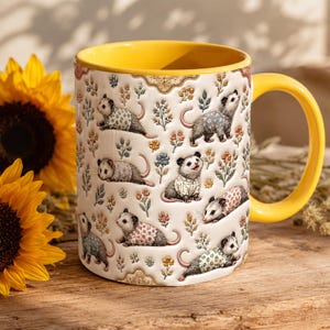 May include: A white ceramic mug with a bright yellow interior and handle, featuring a repeating pattern of small opossums wearing various floral-patterned sweaters, interspersed with delicate illustrations of colourful wildflowers and foliage.