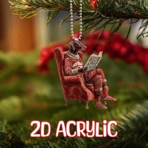 May include: A 2D acrylic Christmas ornament featuring a dog with a brown coat and a sweater, reading a book while seated in a red armchair. The ornament is suspended from a silver chain. The text "2D ACRYLIC" is displayed in red and white.