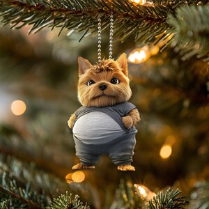 以下が含まれることがあります： 銀色のチェーンにぶら下がっている、茶色と黄褐色のヨークシャーテリアの犬が、灰色の服を着ています。犬は、白いお腹の灰色の服を着ています。犬はカメラを見ています。