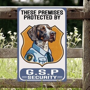May include: A metal sign featuring the text "THESE PREMISES PROTECTED BY" above a dog illustration in a security guard uniform. The sign also displays "G.S.P SECURITY" in a blue banner.