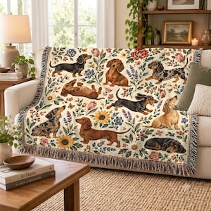 May include: A woven throw blanket featuring a cream background with a repeating pattern of various dachshund dogs in black, tan, and dapple coats, interspersed with colourful illustrations of sunflowers, tulips, and lavender, finished with a fringed edge.