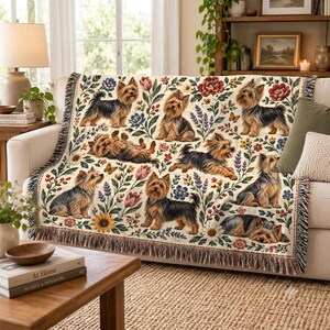 May include: A woven throw blanket featuring a repeating pattern of Yorkshire Terriers in various poses, including sitting, lying down, and running, interspersed with colourful floral illustrations of tulips, sunflowers, and lavender on a cream background.