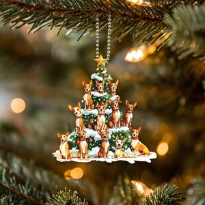 May include: A Christmas tree ornament featuring a group of brown and white dogs of various sizes, all wearing festive red and green collars, gathered around a green Christmas tree decorated with red ornaments and white snow.