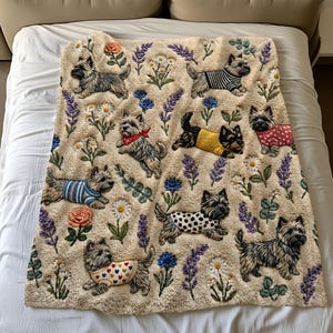 Op de afbeelding: Een crèmekleurige plaid met een herhalend patroon van Cairn Terriers in diverse outfits, waaronder gestreepte shirts, truien en vesten met stippen, afgewisseld met geborduurde bloemen zoals lavendel, madeliefjes en rozen.