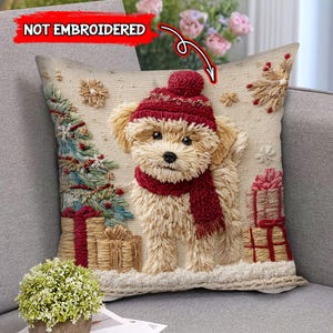 Op de afbeelding: Een beige sierkussen met een terriër puppy met een rode muts en sjaal. De puppy is omringd door een kerstboom, ingepakte cadeaus en sneeuwvlokken. De tekst "NOT EMBROIDERED" staat in de linkerbovenhoek.
