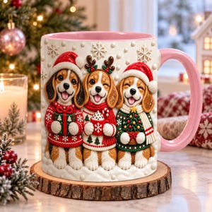 Puede incluir: Una taza de cerámica blanca con interior y asa de color rosa, con un diseño en relieve 3D de tres perros beagle con jerséis y gorros navideños. Los perros sostienen un regalo, una taza de cacao y un pequeño árbol de Navidad decorado.