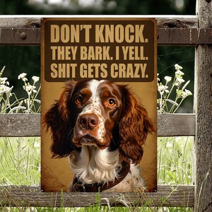 Könnte beinhalten: Ein Metallschild mit dem Text "DON'T KNOCK. THEY BARK. I YELL. SHIT GETS CRAZY." zeigt einen braun-weißen Hund mit Lederhalsband. Das Schild ist an einem Holzzaun mit grasbewachsenem Hintergrund befestigt.