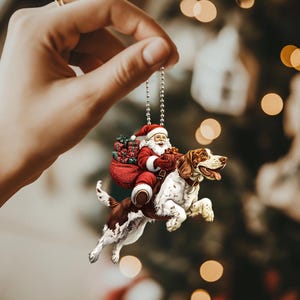 May include: A Christmas ornament featuring a Santa Claus riding a brown and white dog. Santa is wearing a red suit with white trim and a white beard. He is holding a bag of presents and a candy cane. The dog is wearing a red collar and has a brown nose.