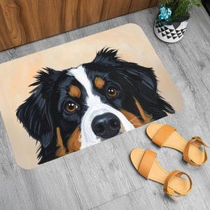 Puede incluir: Una alfombra rectangular con la cara de un perro de montaña bernés, con pelo negro, blanco y marrón. La alfombra está sobre un fondo beige claro. Un par de sandalias color canela están junto a la alfombra sobre un suelo de madera gris.