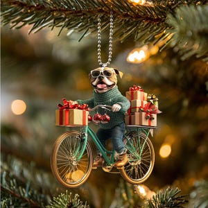 May include: A Christmas tree ornament featuring a bulldog wearing sunglasses and a green sweater, riding a teal bicycle loaded with wrapped gifts. The ornament has a silver chain for hanging and is set against a blurred background of Christmas tree branches and lights.