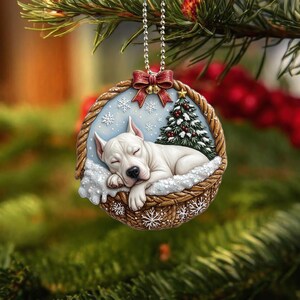 Könnte beinhalten: Weihnachtsschmuck mit einem weißen Hund, der in einem geflochtenen Korb mit Schneeflocken und einem Weihnachtsbaum schläft. Der Schmuck hängt an einer silbernen Kette mit einer roten Schleife.