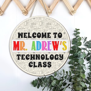 May include: A round wooden sign with the text "WELCOME TO MR. ADREW'S TECHNOLOGY CLASS" in black and colorful letters. The sign has a beige background with a repeating pattern of technology-related doodles. A white rope is attached for hanging.