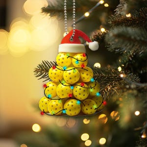 May include: A Christmas tree bauble shaped like a Christmas tree made of yellow pickleball balls with red, blue and green lights. The bauble has a red Santa hat on top.