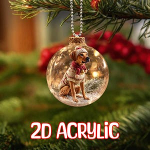 May include: A clear, round Christmas bauble featuring a dog in a hat, scarf, and sweater. The bauble hangs from a silver chain. The text "2D ACRYLIC" is displayed in red and white at the bottom of the image.