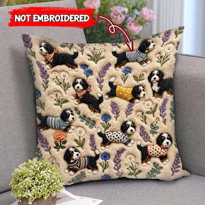 May include: Beige decorative pillow featuring a repeating pattern of black, white, and brown dogs wearing various outfits. The pillow also has floral and plant designs in blue, purple, and white. The text "NOT EMBROIDERED" is in the upper left corner.
