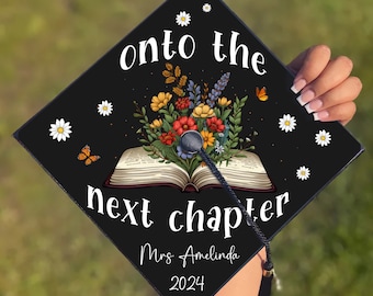 May include: Black graduation cap with a white tassel. The cap features a book with flowers and butterflies, and the text "onto the next chapter" and "Mrs. Amelinda 2024".