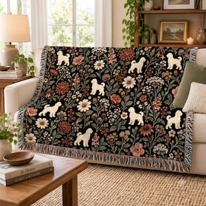 May include: A woven throw blanket featuring a repeating pattern of cream-coloured curly-haired dogs interspersed with various wildflowers in shades of red, orange, and white against a black background, finished with a decorative fringed edge.