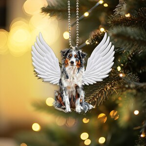 Può includere: Un ornamento decorativo raffigurante un cane pastore australiano con ali d'angelo bianche. Il cane ha un mantello multicolore nero, bianco e marrone. L'ornamento è appeso a una catena d'argento su uno sfondo sfocato di luci natalizie.