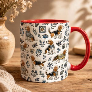 Könnte beinhalten: Ein weißer Keramikbecher mit leuchtend rotem Innenraum und Henkel, verziert mit einem wiederkehrenden Muster aus Beagle-Hunden in verschiedenen Posen, einige tragen Pullover, durchsetzt mit blauen Blumenmotiven, kleinen Herzen und Paisleymustern.