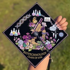 May include: Black graduation cap decorated with a gold microscope, flowers, and scientific illustrations. The cap features the text "Biotechnology Class of 2026" in gold lettering, with a gold and black border.