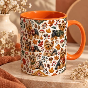 May include: A ceramic mug with an orange handle and interior, featuring a repeating pattern of Yorkshire Terriers wearing jumpers, surrounded by autumn leaves, acorns, berries, and mushrooms on a white background.