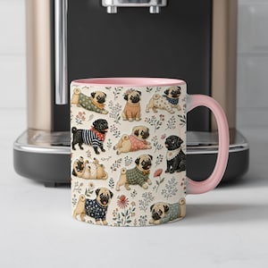 May include: A ceramic mug with a light pink handle and interior, featuring a repeating pattern of various pug dogs wearing sweaters and bandanas, interspersed with small colorful floral illustrations on a cream-colored background.