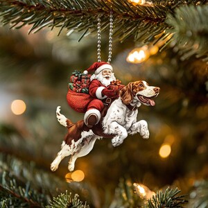 May include: A Christmas ornament featuring a Santa Claus riding a brown and white dog. Santa is wearing a red suit and holding a bag of presents. The dog is wearing a red collar.