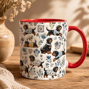 May include: A white ceramic mug with a bright red interior and handle, featuring a repeating pattern of Rottweiler dogs wearing patterned sweaters, interspersed with blue floral motifs, small hearts, and dragonfly illustrations.
