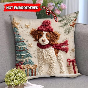 May include: Embroidered Christmas throw pillow featuring a dog wearing a red hat and scarf. The dog is standing next to a Christmas tree and presents. The pillow has a beige background with a festive design.