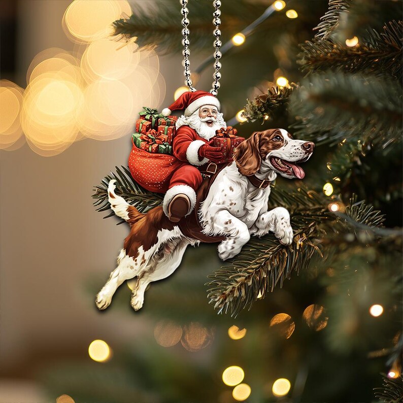 May include: A Christmas ornament featuring a Santa Claus riding a brown and white dog. Santa is wearing a red suit with white trim and a white beard. He is holding a bag of presents and a red gift box. The dog is wearing a red collar and has a brown nose.