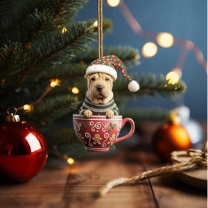 May include: A Shar Pei dog ornament wearing a Christmas sweater and hat, sitting in a red teacup. The teacup is decorated with gingerbread men and holly. The ornament hangs from a string in front of a Christmas tree.