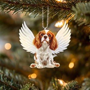 Könnte beinhalten: Ein weißer Keramik-Anhänger mit einem Cavalier King Charles Spaniel mit braunem und weißem Fell, der Engelsflügel trägt. Der Hund sitzt mit leicht geneigtem Kopf. Der Anhänger hängt an einer silbernen Kette.