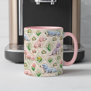Puede incluir: Una taza de cerámica con interior y asa de color rosa claro, que presenta un patrón repetitivo de rombos acolchados en el exterior. El diseño incluye ilustraciones de perros pequeños con cuadros azules y rosas, intercaladas con varios motivos florales coloridos.