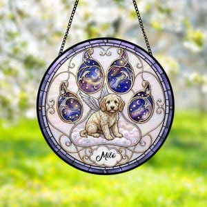 Könnte beinhalten: Ein runder Buntglas-Suncatcher mit einem Pfotenabdruck-Design, in dessen Mitte ein cremefarbener Welpe mit Engelsflügeln auf Wolken sitzt. Die Pfotenballen zeigen dunkelblaue Galaxienmuster mit Sternen und Monden. Der Name Milo ist in Schreibschrift zu sehen.