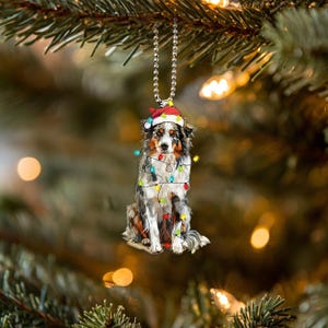 May include: A blue merle Australian Shepherd wearing a Santa hat is tangled in Christmas lights. The dog is sitting and looking at the camera. The ornament is ideal for a Christmas tree.