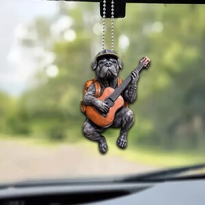 May include: A black dog figurine wearing a hat and a backpack, playing a brown acoustic guitar. The figurine is hanging from a silver chain.