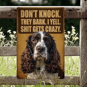 Könnte beinhalten: Ein rechteckiges Metallschild mit dem Text "DON'T KNOCK. THEY BARK. I YELL. SHIT GETS CRAZY." zeigt einen braun-weißen Hund. Das Schild ist an einem Holzzaun befestigt, mit grünem Gras und weißen Blumen im Hintergrund.