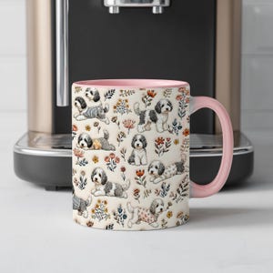 Op de afbeelding: Een keramische mok met een lichtroze handvat en binnenkant, voorzien van een all-over patroon van kleine zwart-witte honden met krullend haar in diverse houdingen, afgewisseld met kleurrijke bloemenillustraties op een crèmekleurige achtergrond.