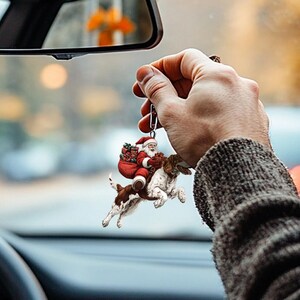 May include: A red and white Santa Claus riding a brown and white dog with a bag of presents. The ornament is hanging from a rearview mirror in a car.